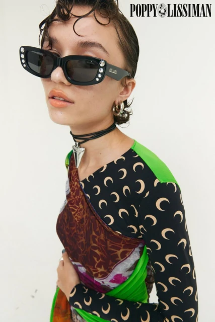 Woman wearing black sunglasses with jeweled accents and a printed top poses against a white backdrop in a studio setting.